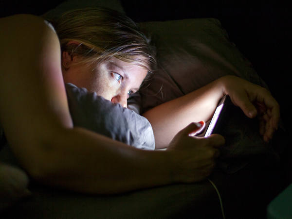 Eine junge Frau liegt im Bett und hat ihr Blick auf das leuchtende Smarphone gerichtet.