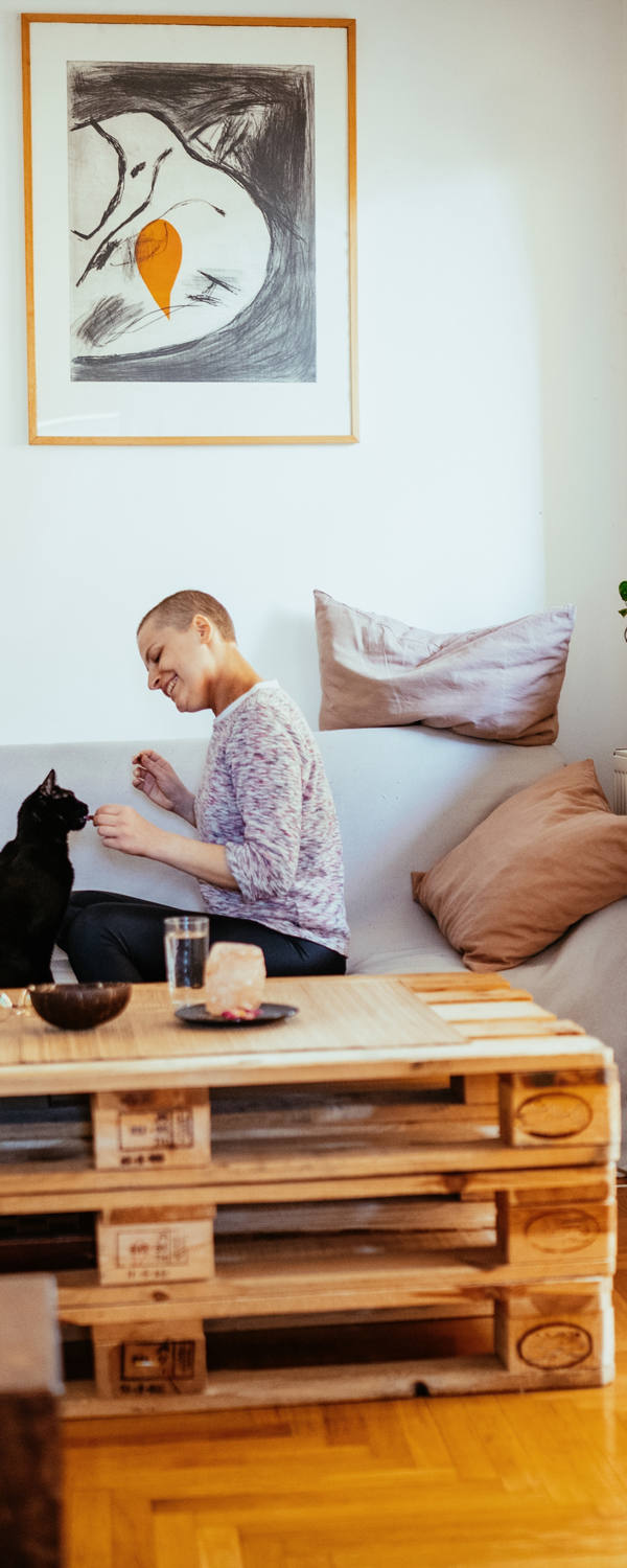 Eine Frau mit fast keinen Haaren und ihr Partner sitzen auf dem Sofa im Wohnzimmer und spielen glücklich mit der Katze.