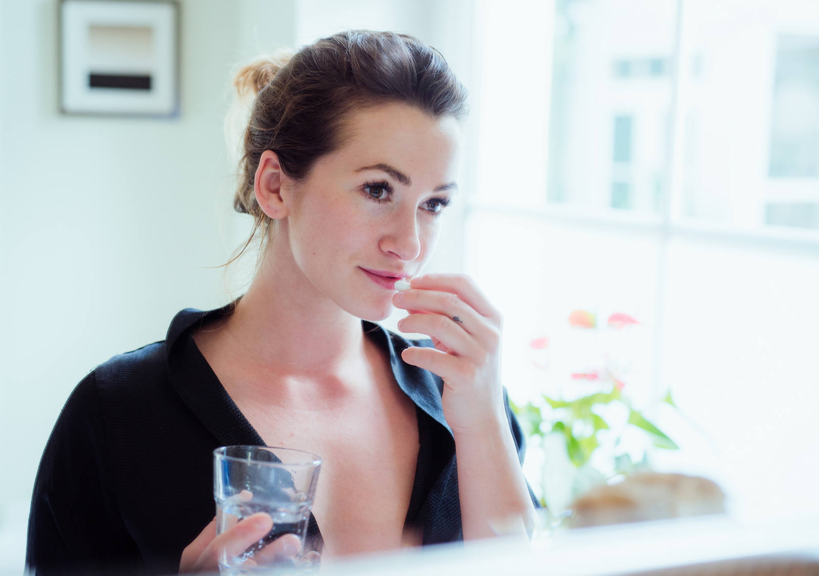 A woman stands in front of a mirror with a glass of water in her hand and takes her food supplements.