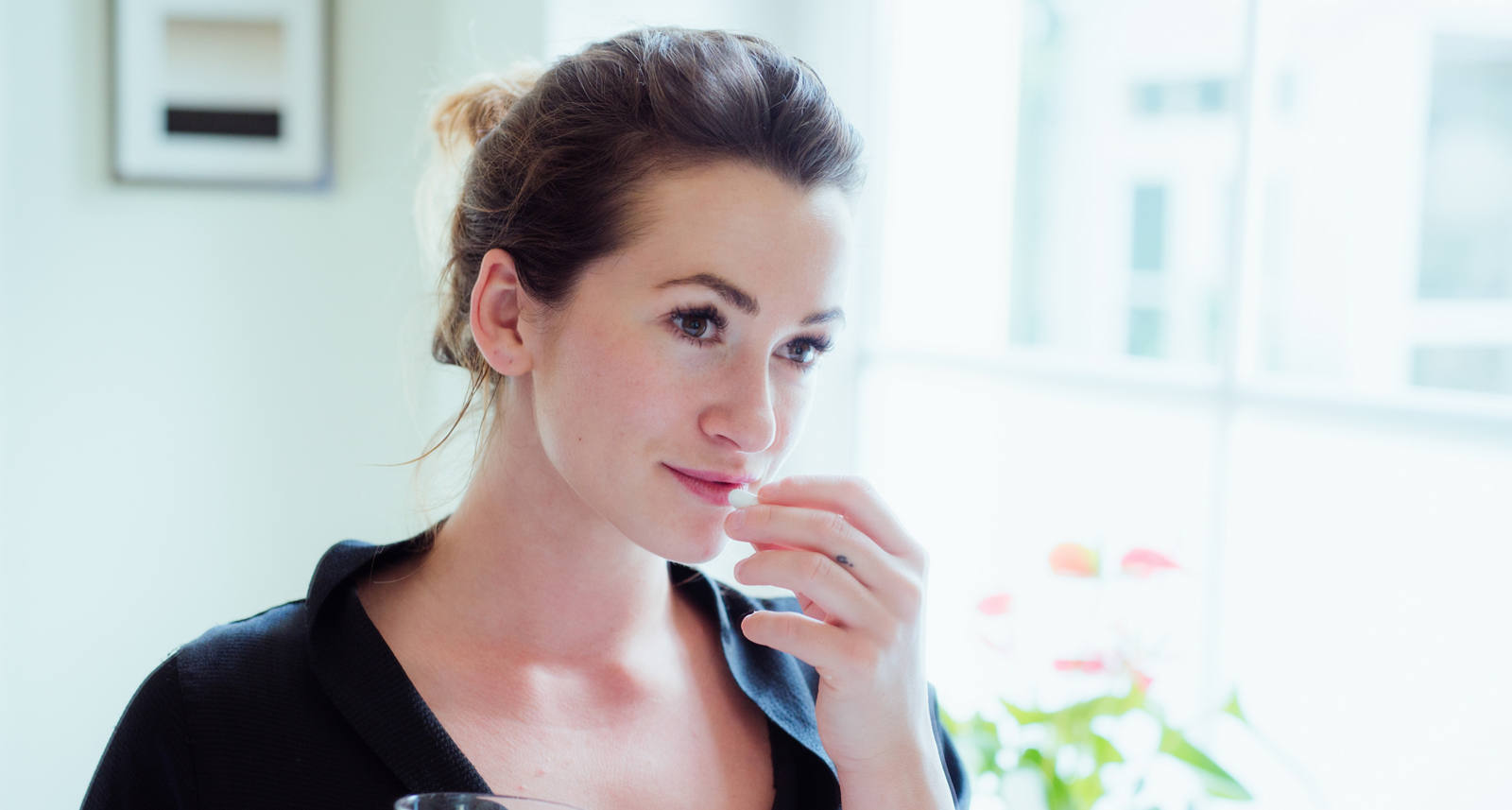 A woman stands in front of a mirror with a glass of water in her hand and takes her food supplements.