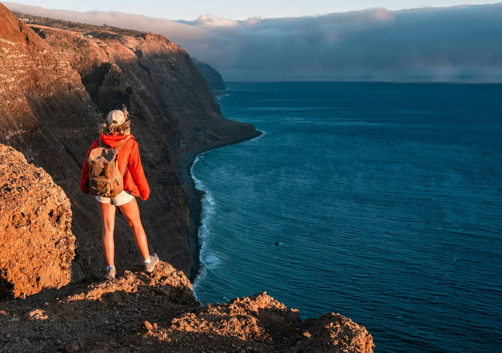 Young woman with backpack looking down from cliffs on sea bay
