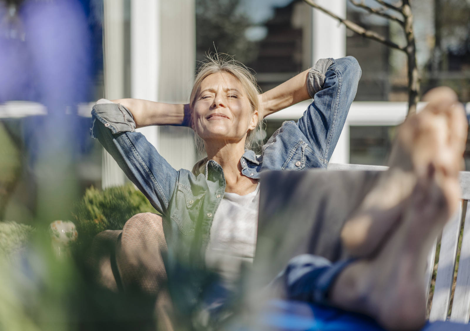 An older woman lies relaxed on a bench in the garden, enjoying the sun’s rays with her eyes closed.