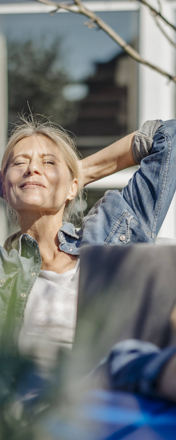An older woman lies relaxed on a bench in the garden, enjoying the sun’s rays with her eyes closed.