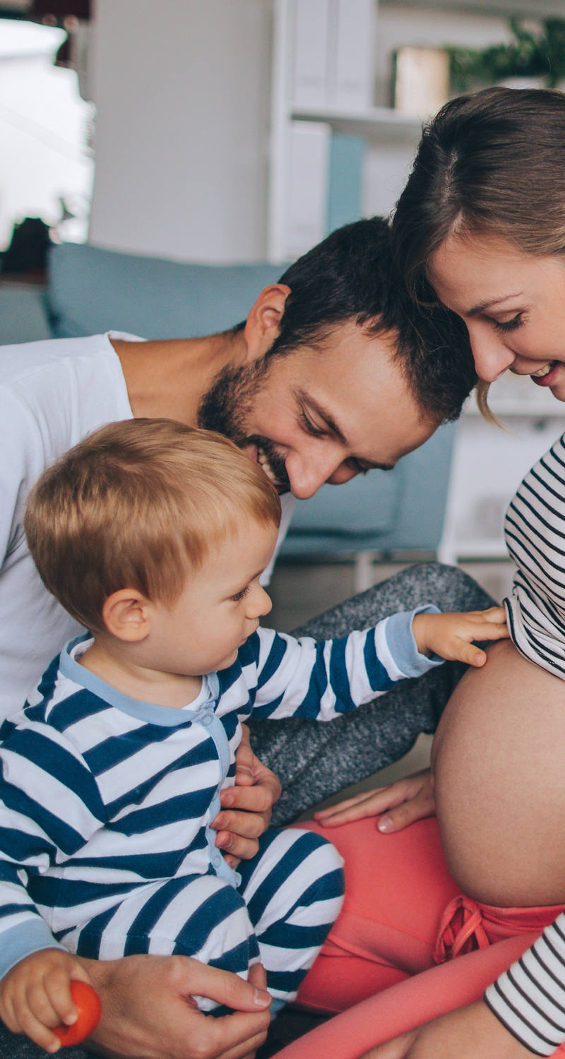 Eine schwangere Frau sitzt mit ihrem Partner und Kind auf dem Boden, während das Kind sanft über ihren Bauch streicht