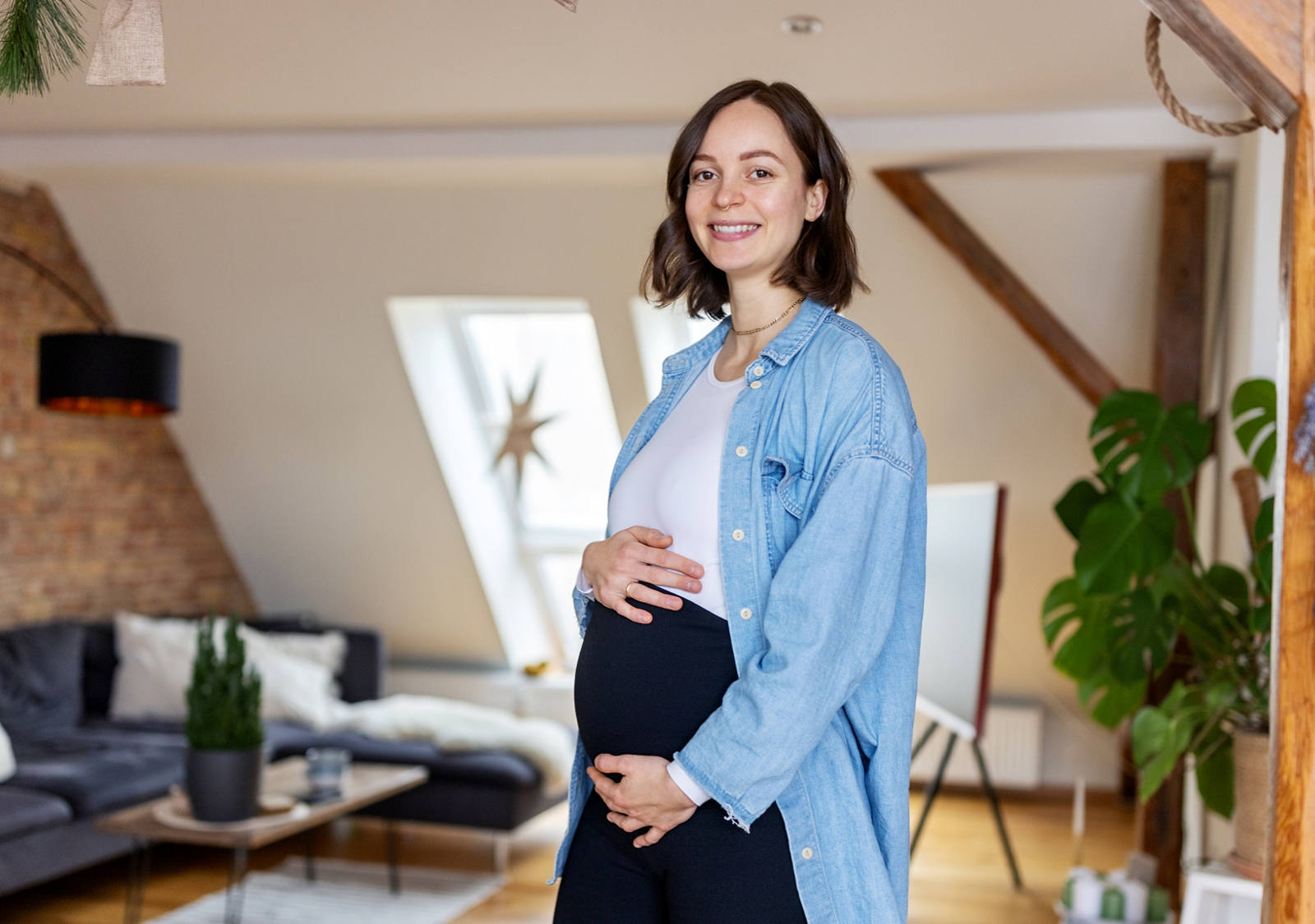 Schwangere Frau steht im Wohnzimmer und zeigt ihren Babybauch und strahlt in die Kamera.