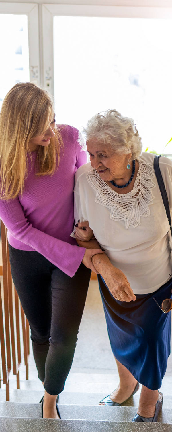 An older woman is supported by another woman by her arm while both walk up the stairs in the staircase.