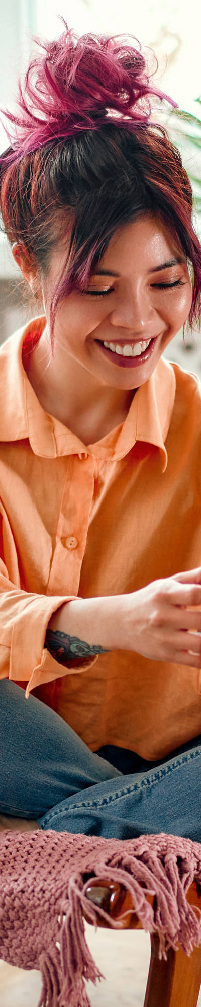 Junge asiatische Frau sitzt auf Stuhl im Wohnzimmer und bedient grinsend ihr Smartphone.