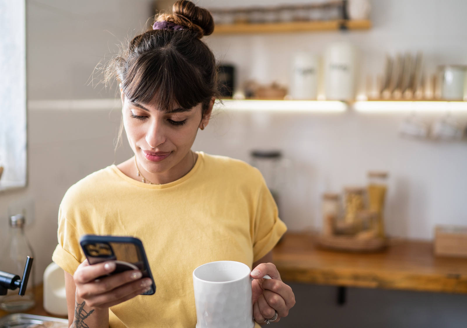 Una donna con una maglietta gialla è in cucina e guarda lo smartphone mentre tiene in mano una tazza di caffè.