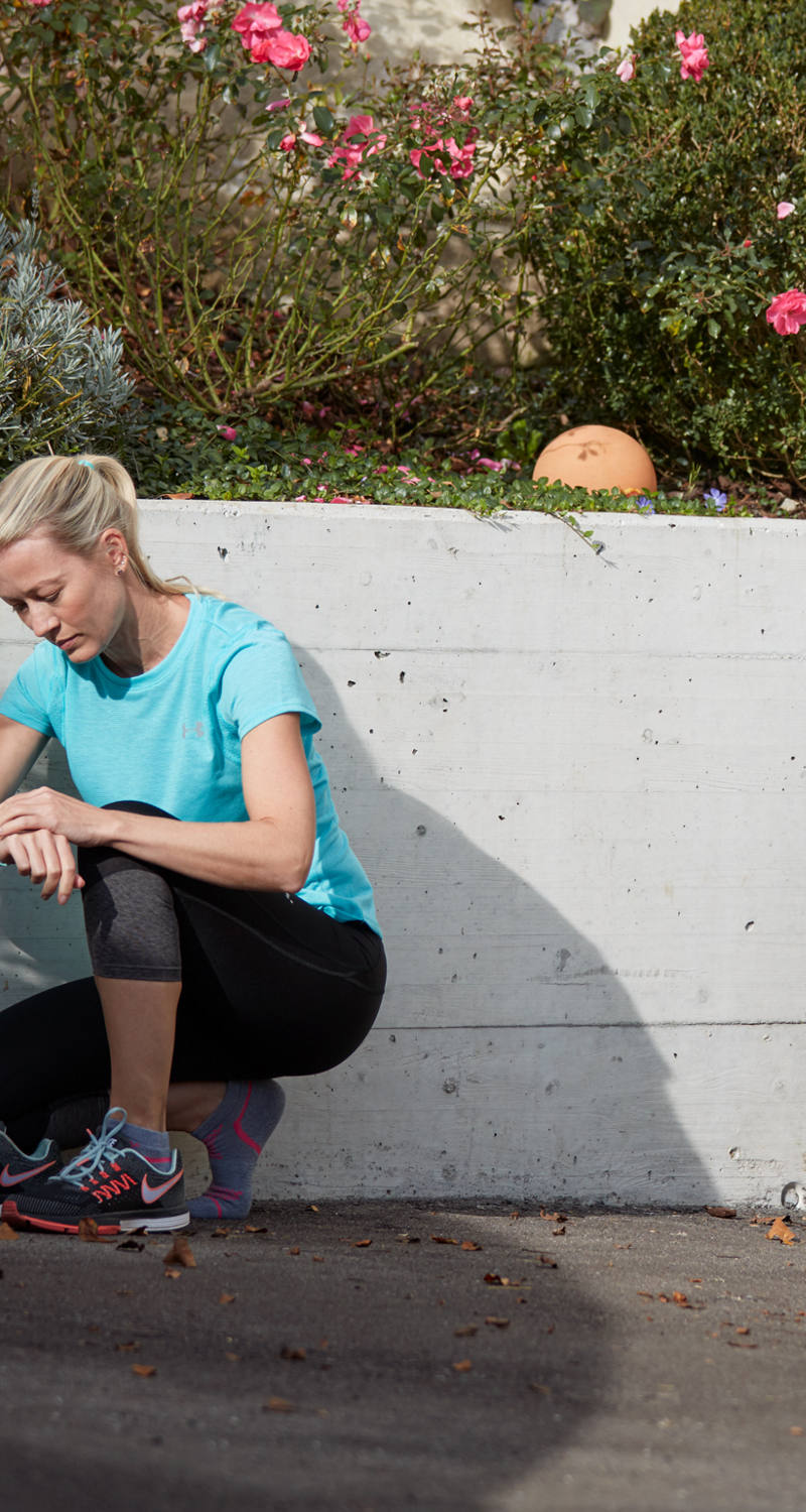 Dehors, sur l’esplanade, une femme en tenue de sport enfile ses chaussures de sport et examine sa montre connectée.