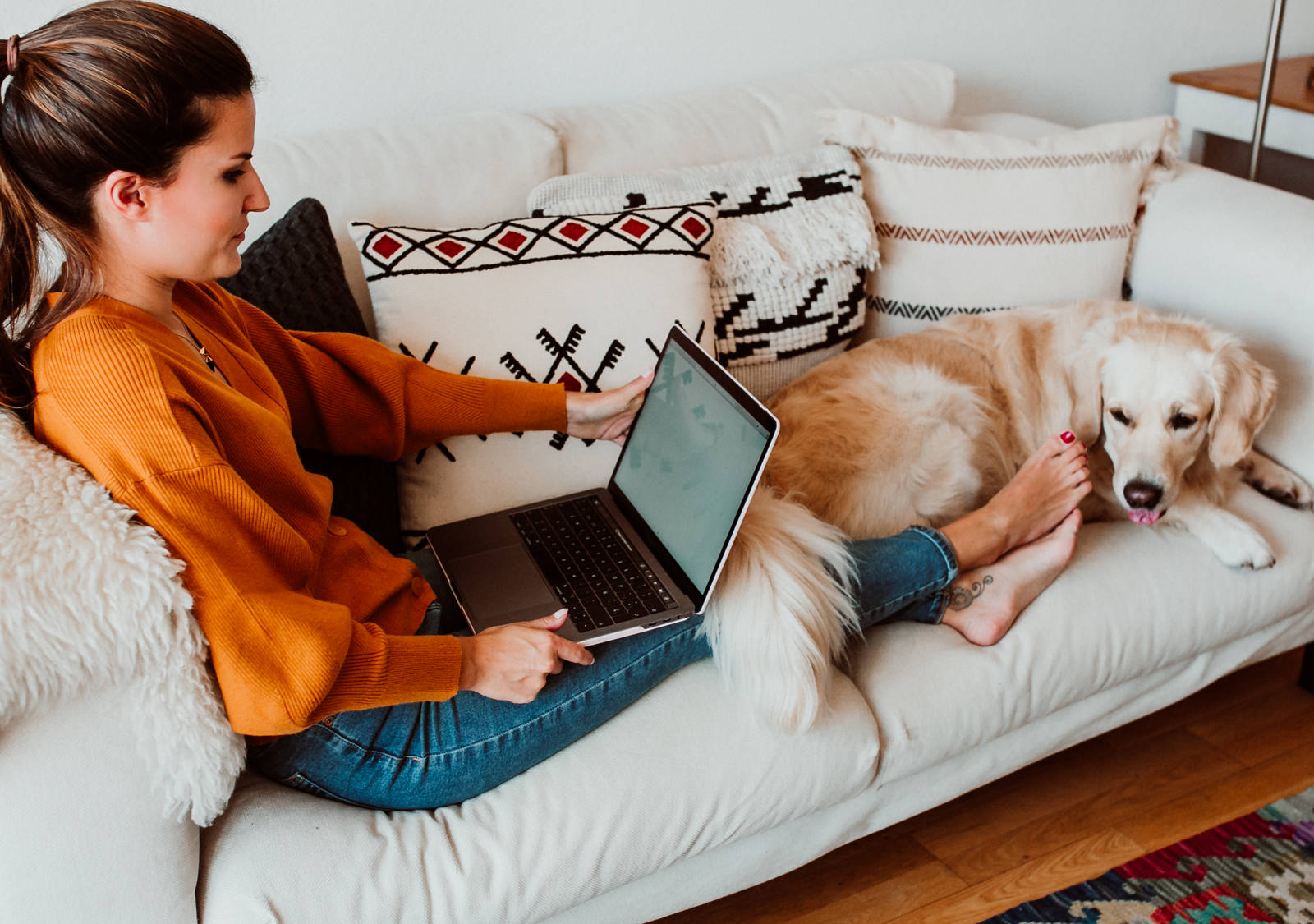 Woman sits on a white sofa with her legs outstretched and bare feet working on a laptop. At her feet lies a light brown Golden Retriever dog.