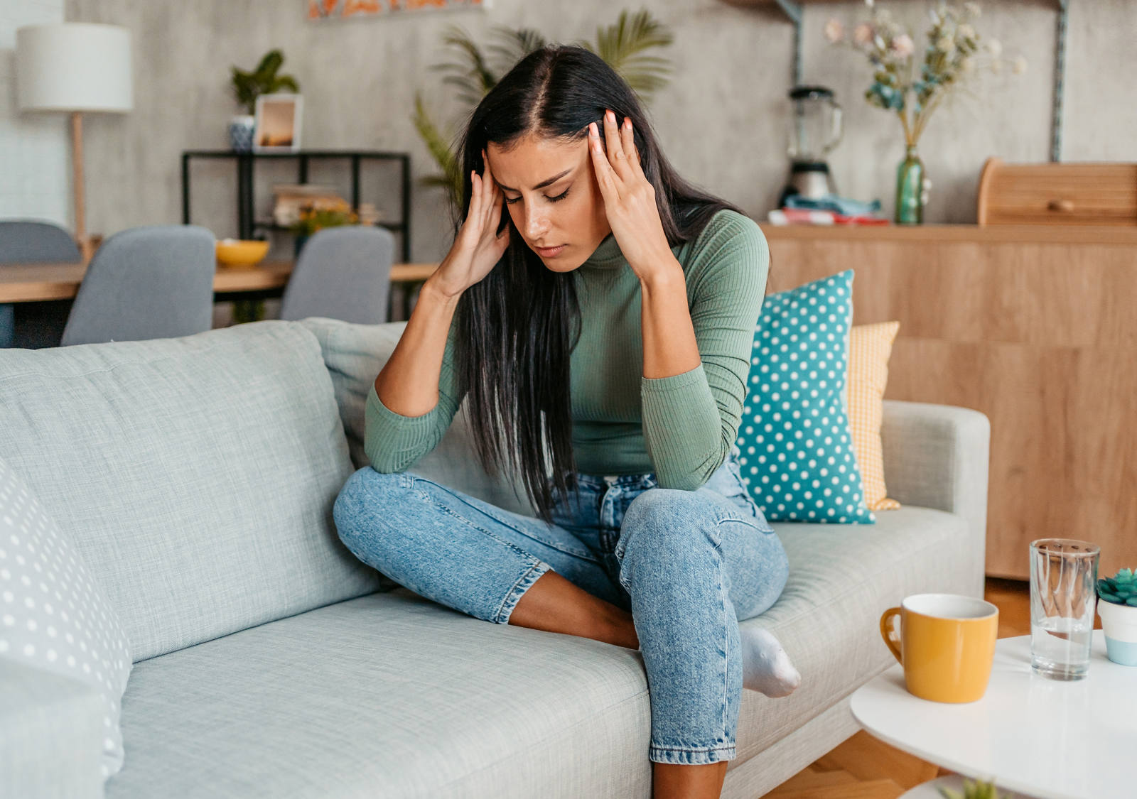 A woman sits on the sofa with her eyes closed, holding her temples and suffering from a headache.