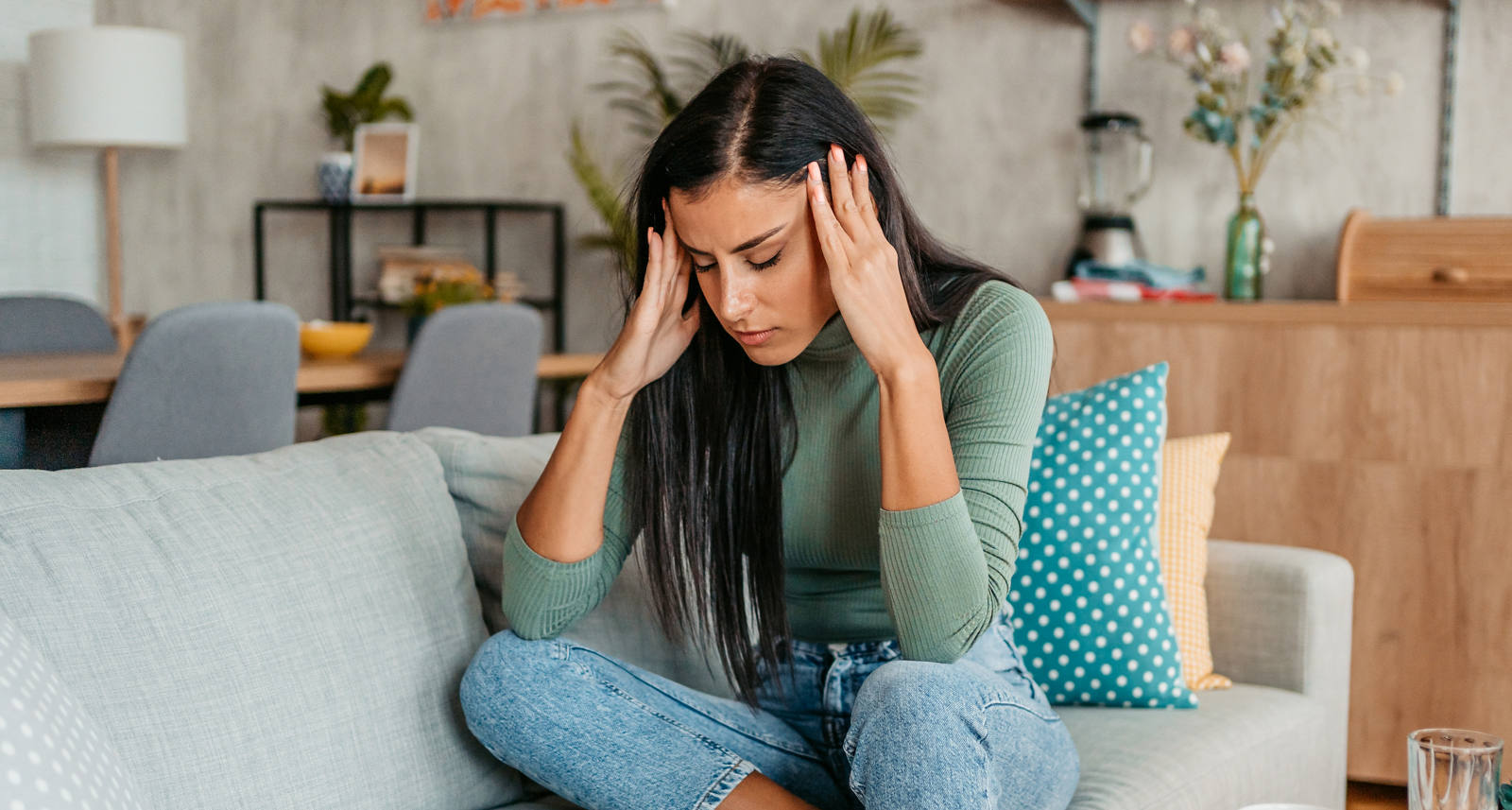 A woman sits on the sofa with her eyes closed, holding her temples and suffering from a headache.