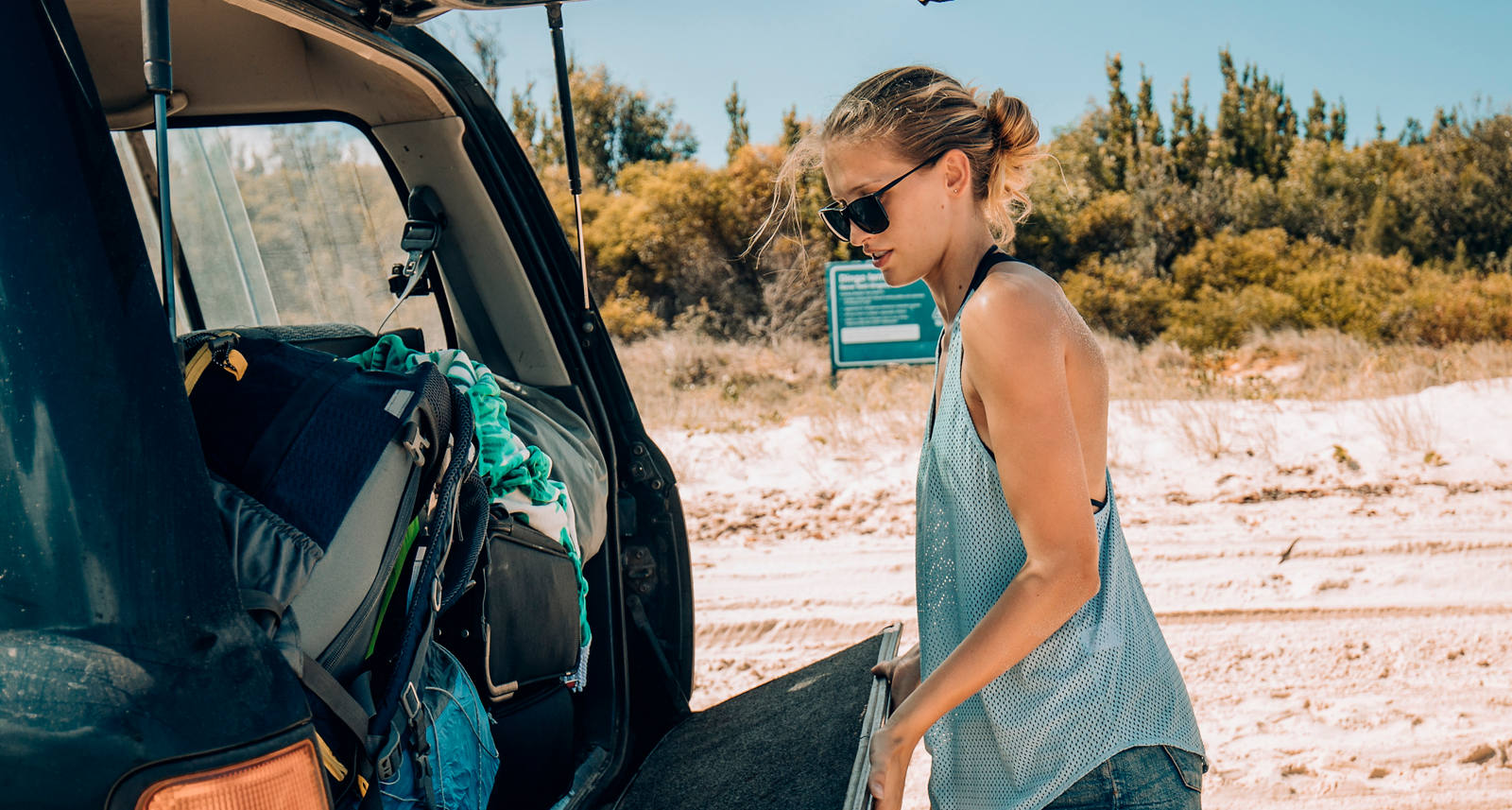 Donna in abito estivo chiude il bagagliaio di un’auto che si trova su una spiaggia.