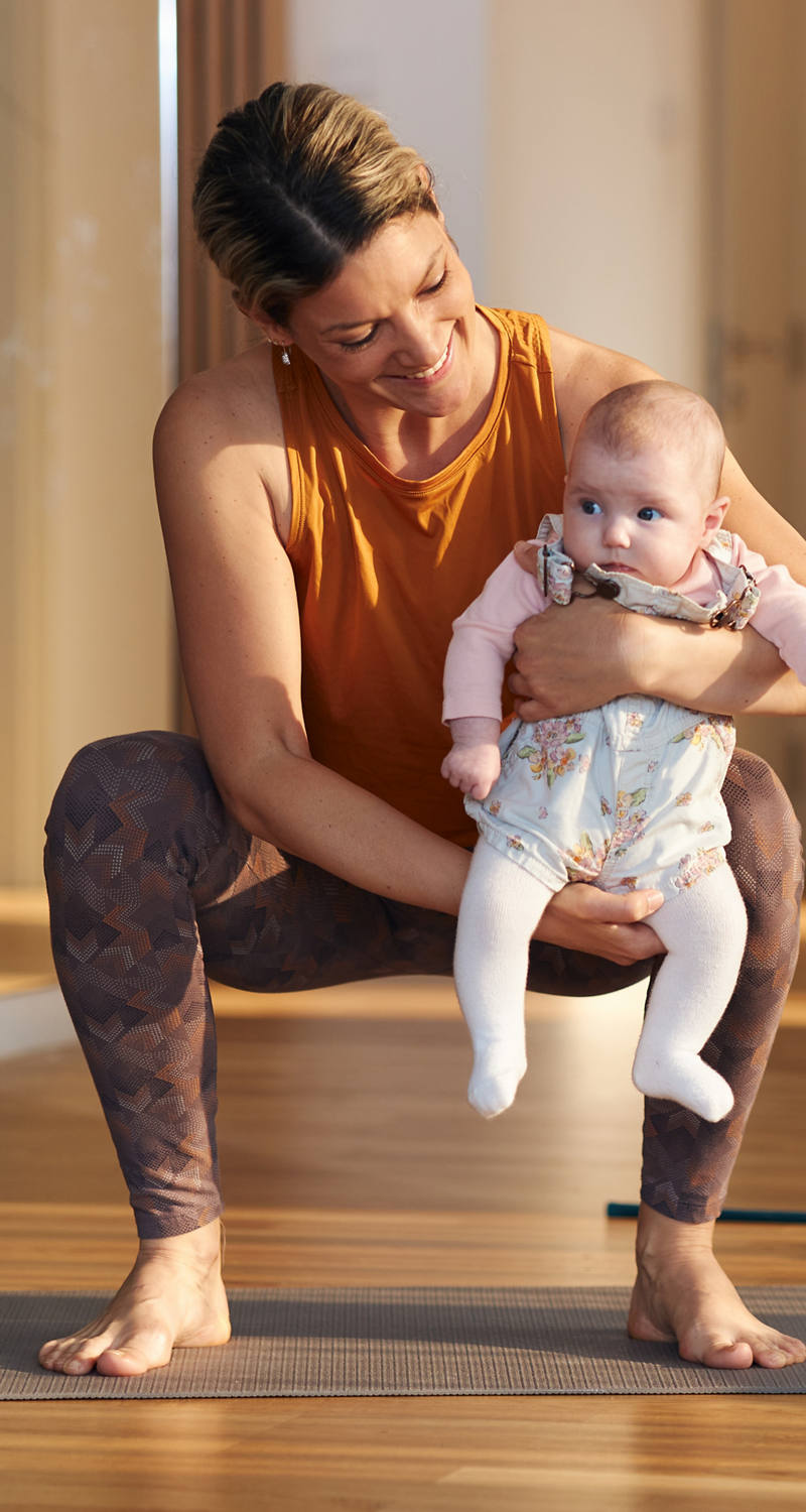 Eine Frau macht Übungen auf einer Sportmatte und hält dabei lachend ihr Baby