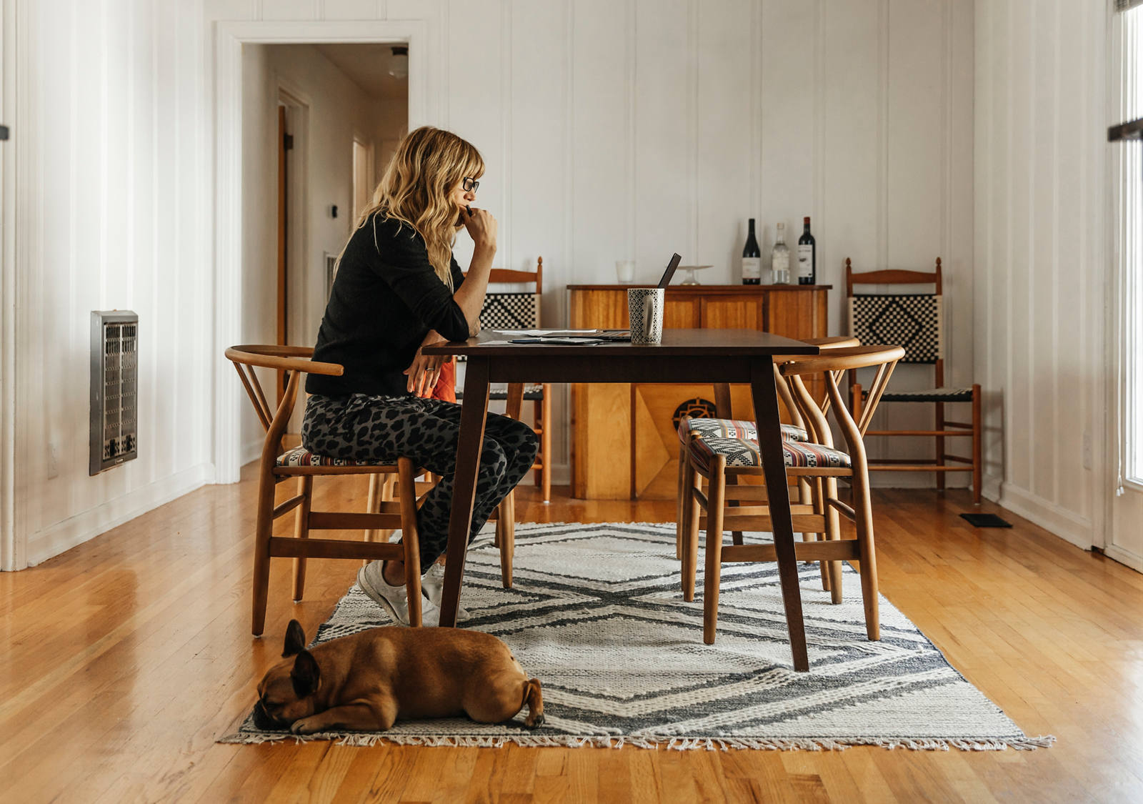 Une femme est assise à la table du salon et étudie son ordinateur portable. Un chien dort à ses pieds.