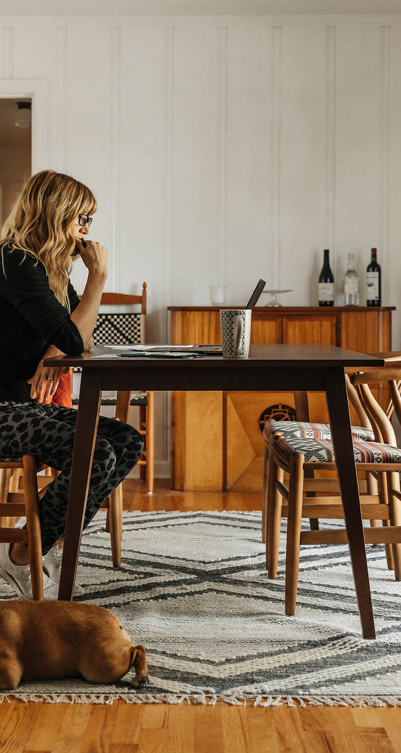 Une femme est assise à la table du salon et étudie son ordinateur portable. Un chien dort à ses pieds.