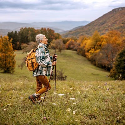Eine ältere Frau wandert mit ihren Wanderstöcken über eine Hügel-Landschaft mit Wald