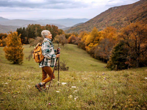 Eine ältere Frau wandert mit ihren Wanderstöcken über eine Hügel-Landschaft mit Wald