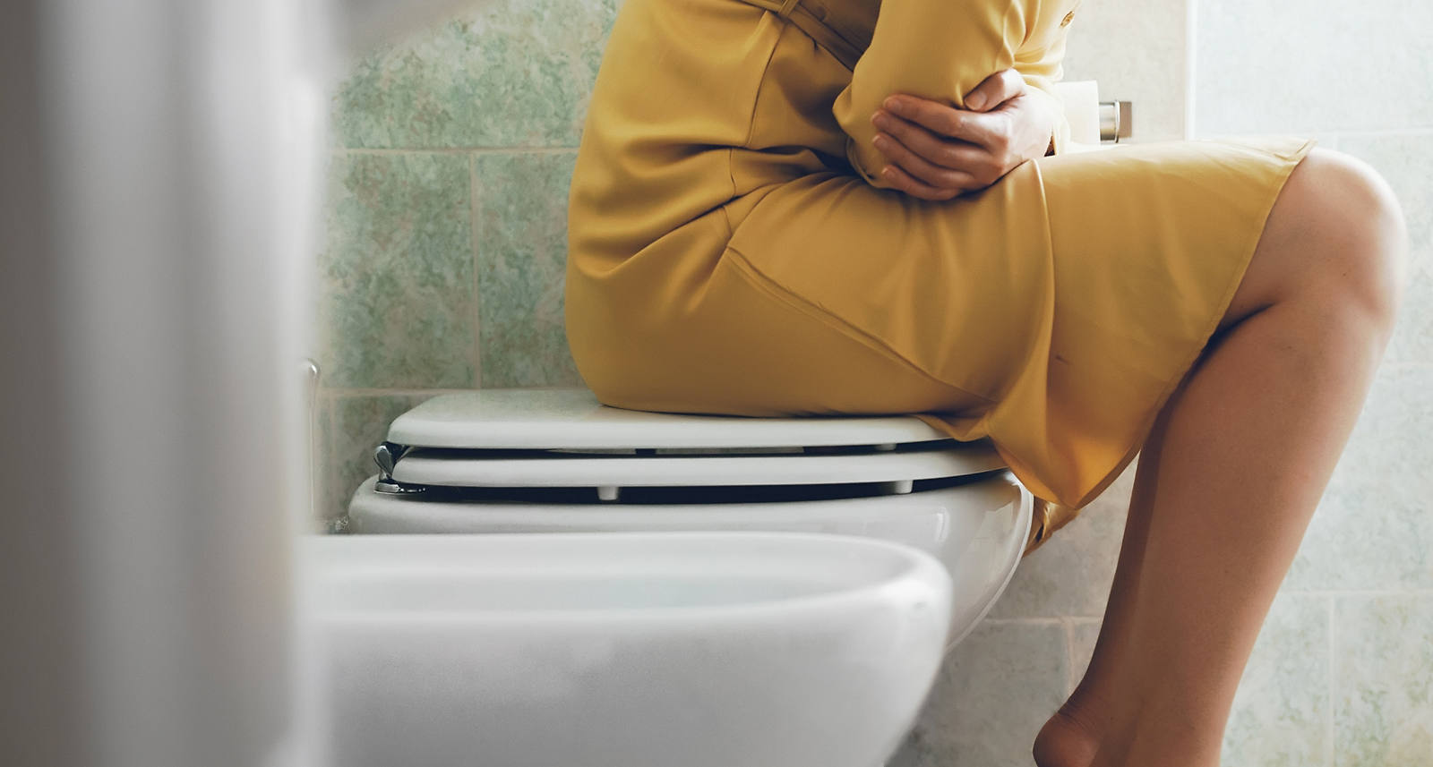 Frau im gelben Kleid fühlt sich unwohl und sitzt auf dem Deckel der Toiletten-Schüssel.