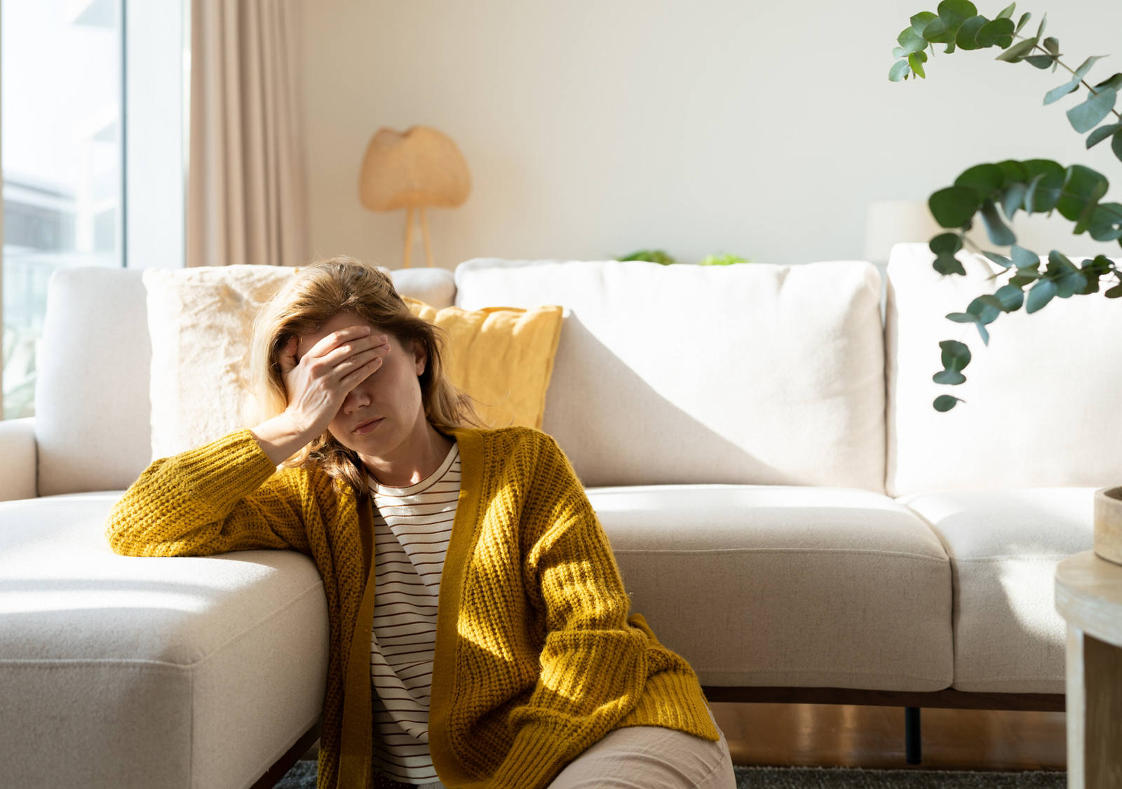 A tired woman sits on the floor in front of the sofa, touching her forehead exhausted.