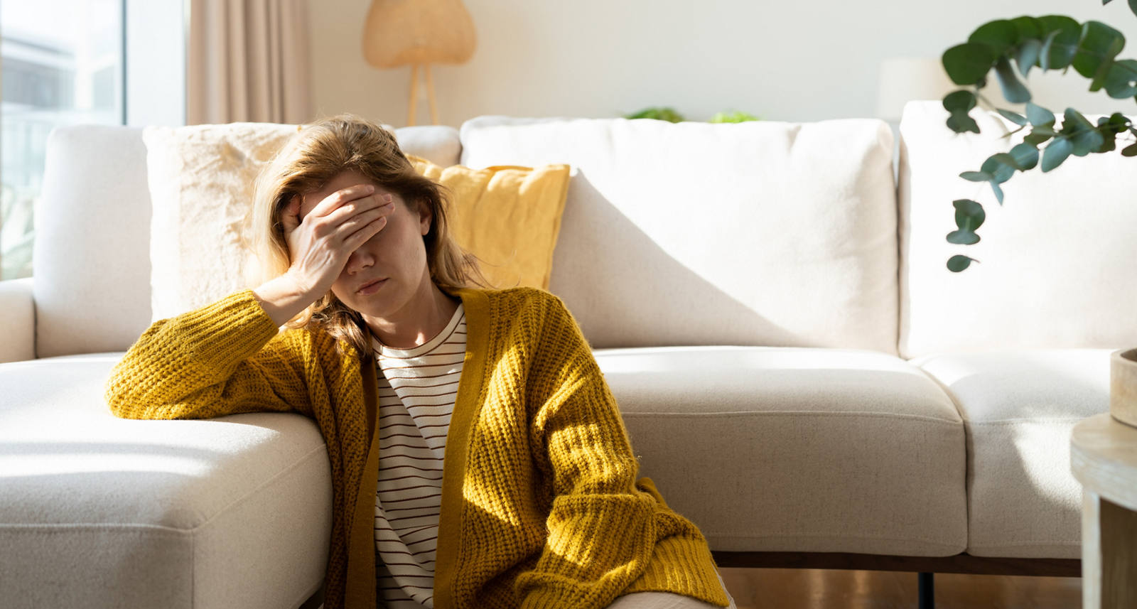 Una donna stanca è seduta per terra davanti al divano e si tocca la fronte, esausta.