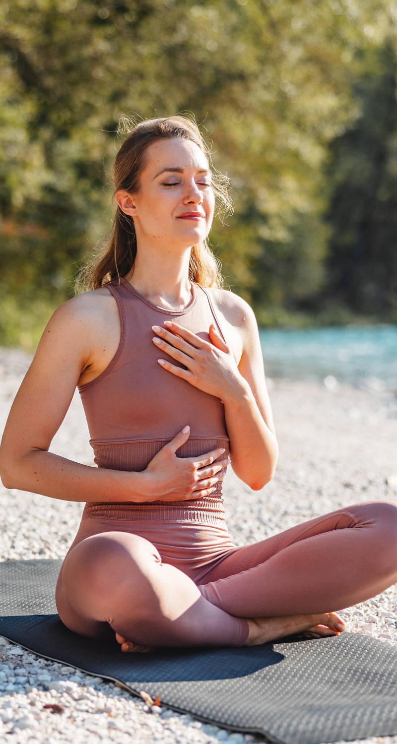 Eine Frau sitzt im Schneidersitz auf einer Yogamatte am Wasser und macht Atemübungen