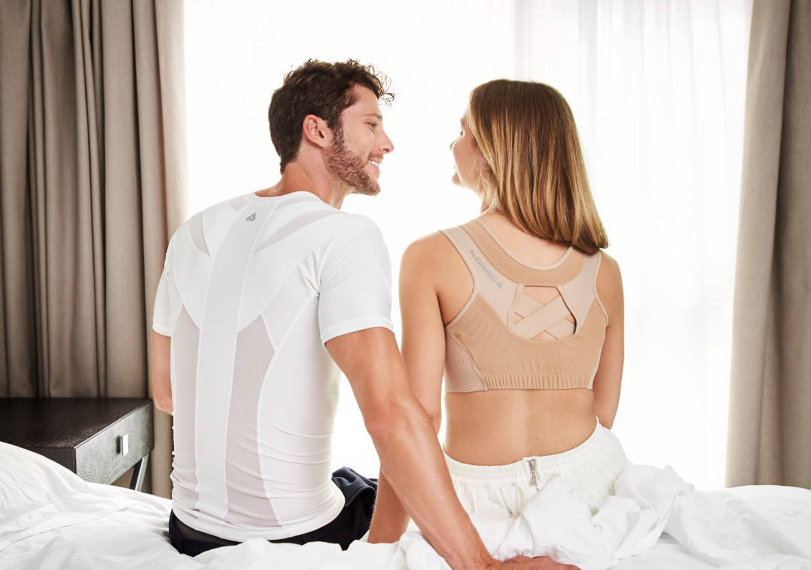 A couple is sitting on the edge of the bed and smiling at each other. Both are wearing a posture shirt.