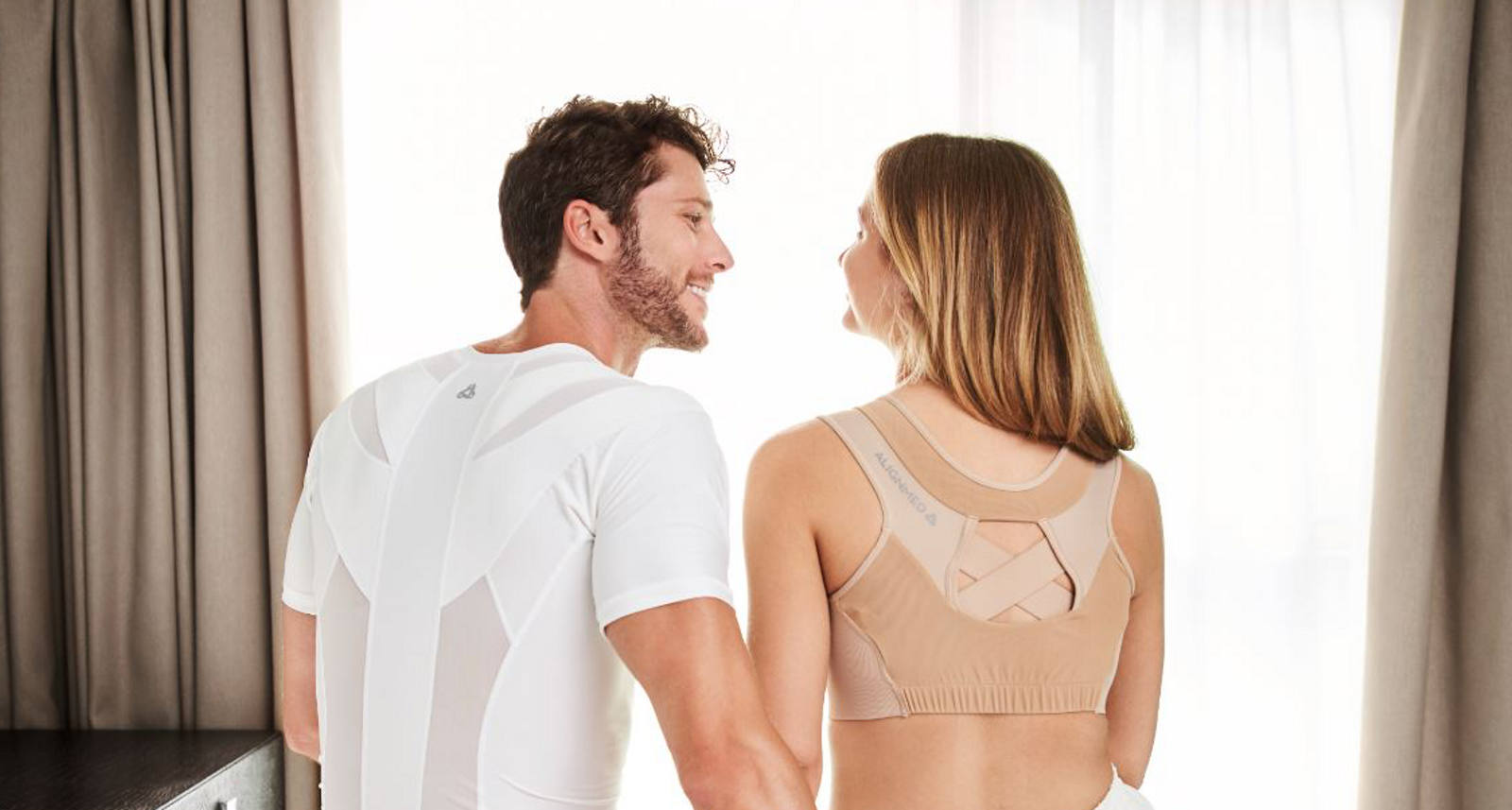 Ein Paar sitzt auf der Bettkante und lächelt sich an. Beide tragen ein Haltungsshirt.