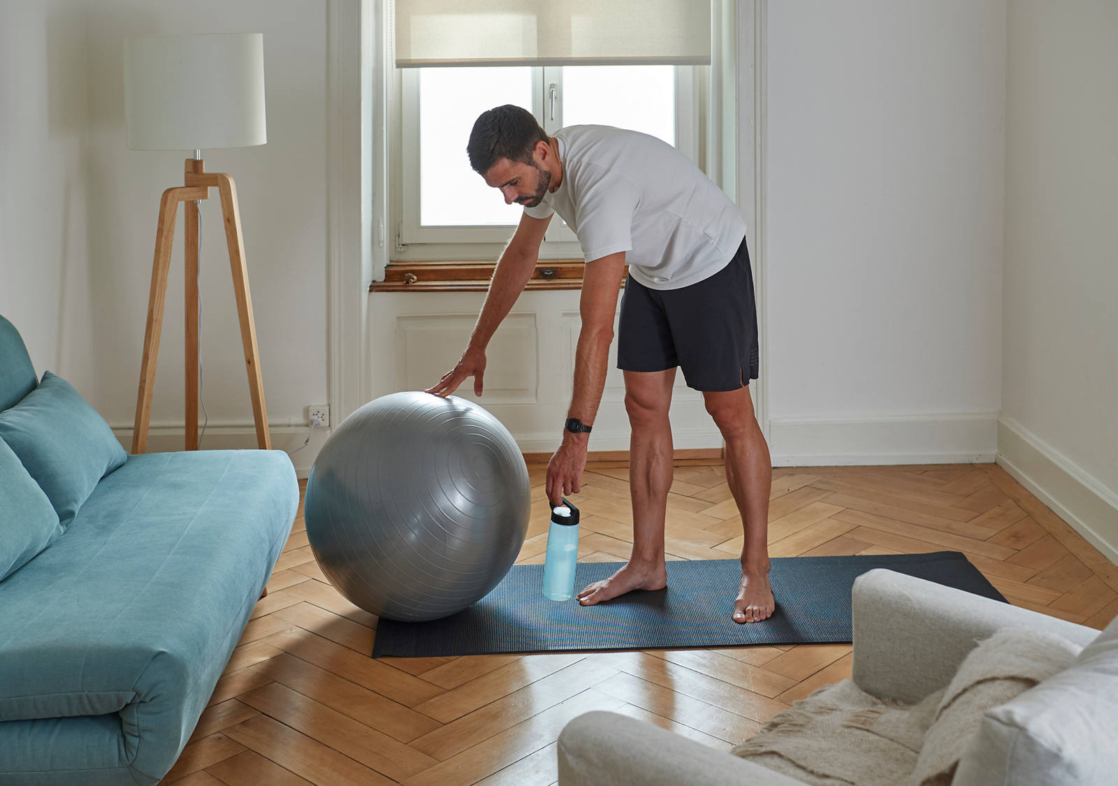 Ein Mann im Wohnzimmer steht auf einer Gymnastikmatte und greift zum Gymnastikball.