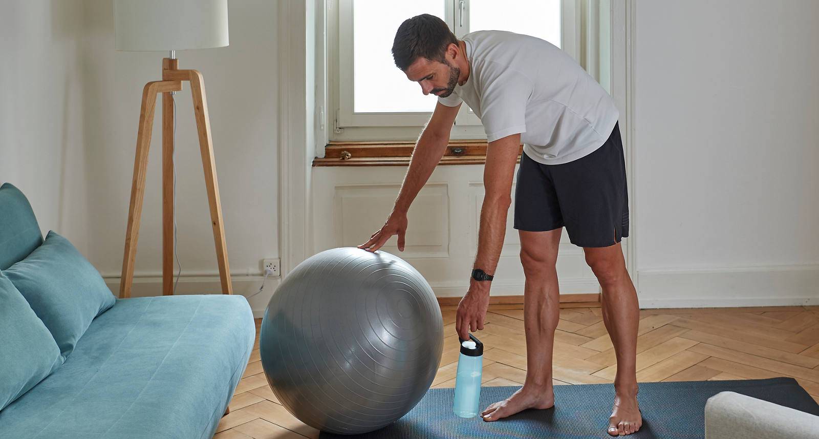 A man in the living room stands on a gymnastics mat and grabs the gymnastics ball.