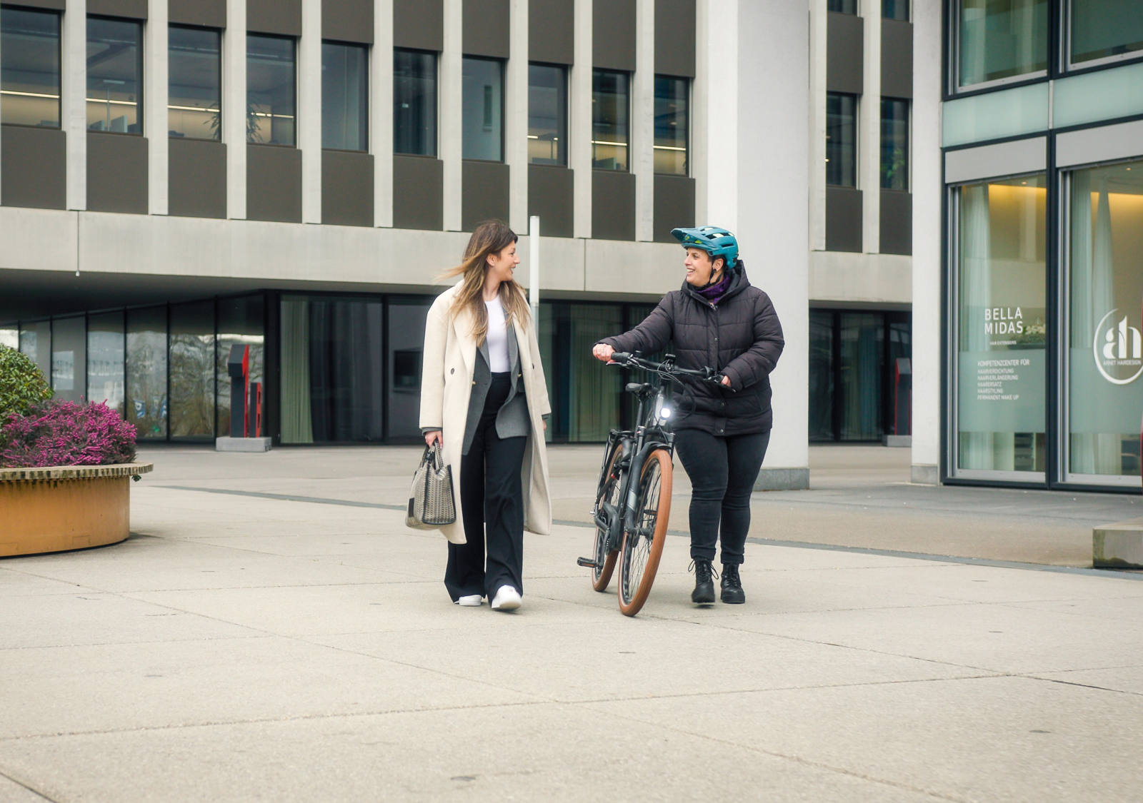 Alla fine della giornata di lavoro, due collaboratrici tornano a casa insieme; una cammina a piedi e l’altra spinge la bicicletta accanto a sé. 