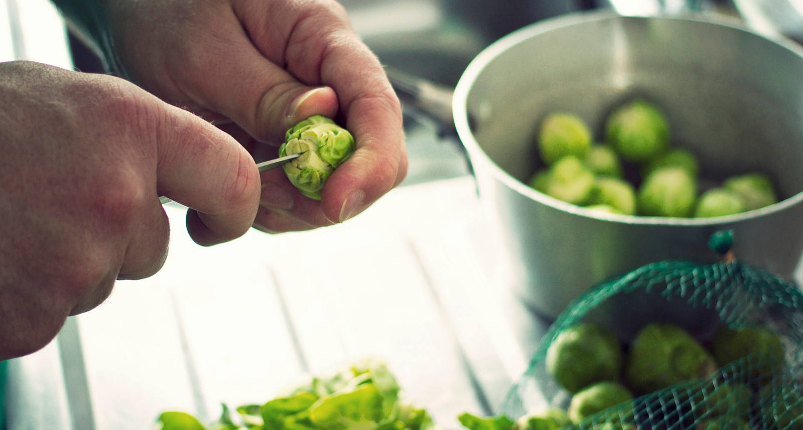 A man in the kitchen has washed a whole net of Brussels sprouts and is now preparing them with a knife