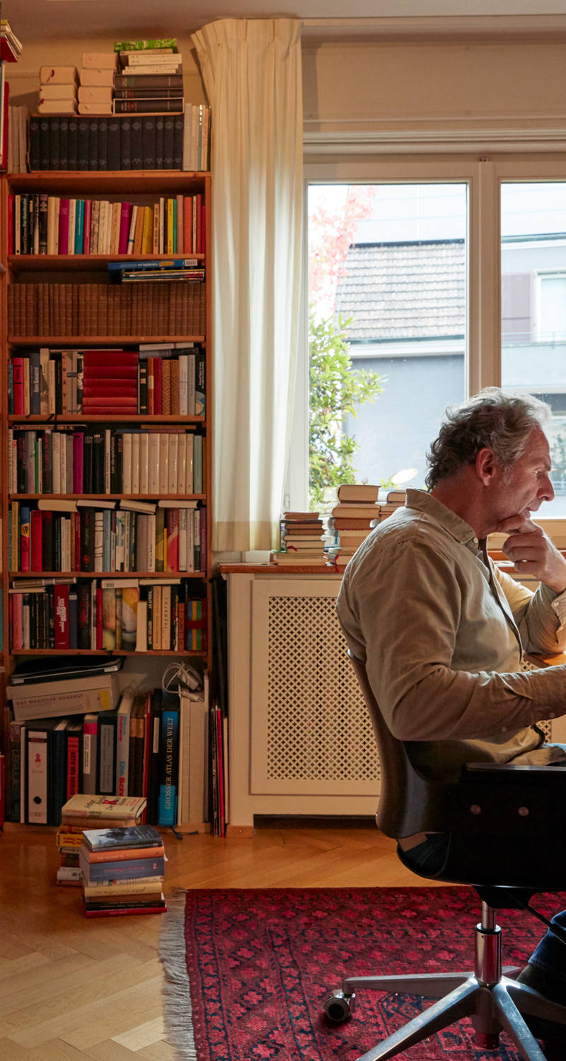 Mann studiert Buch in seinem Büro zu Hause. Im Hintergrund ist eine riesige Bücherwand.