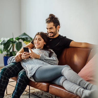 Una coppia giovane si siede sul divano e guarda felicemente smartphone.