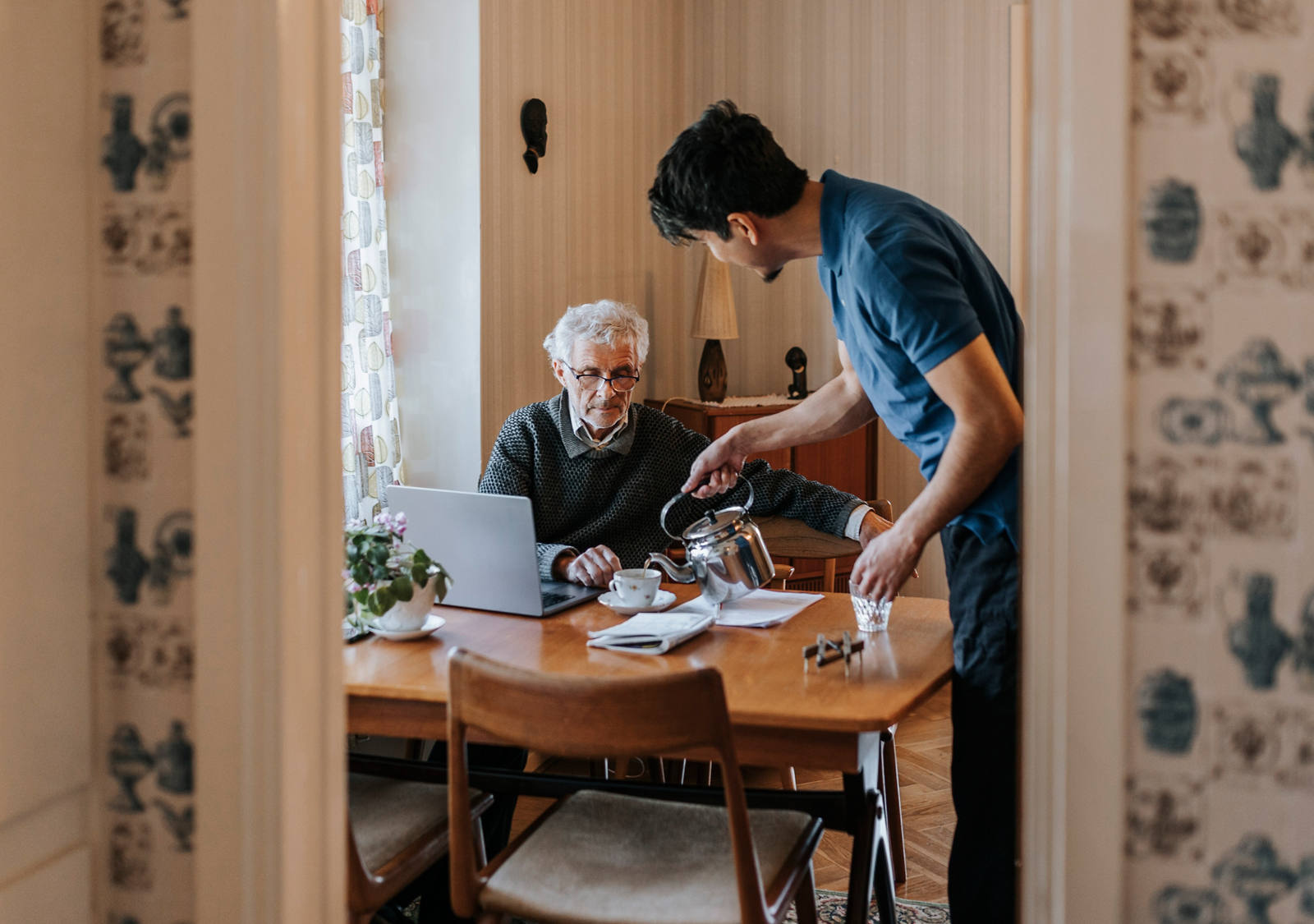 Ein junger Angehöriger hilft einem älteren Herrn zuhause und giesst ihm eine Tasse Tee ein.