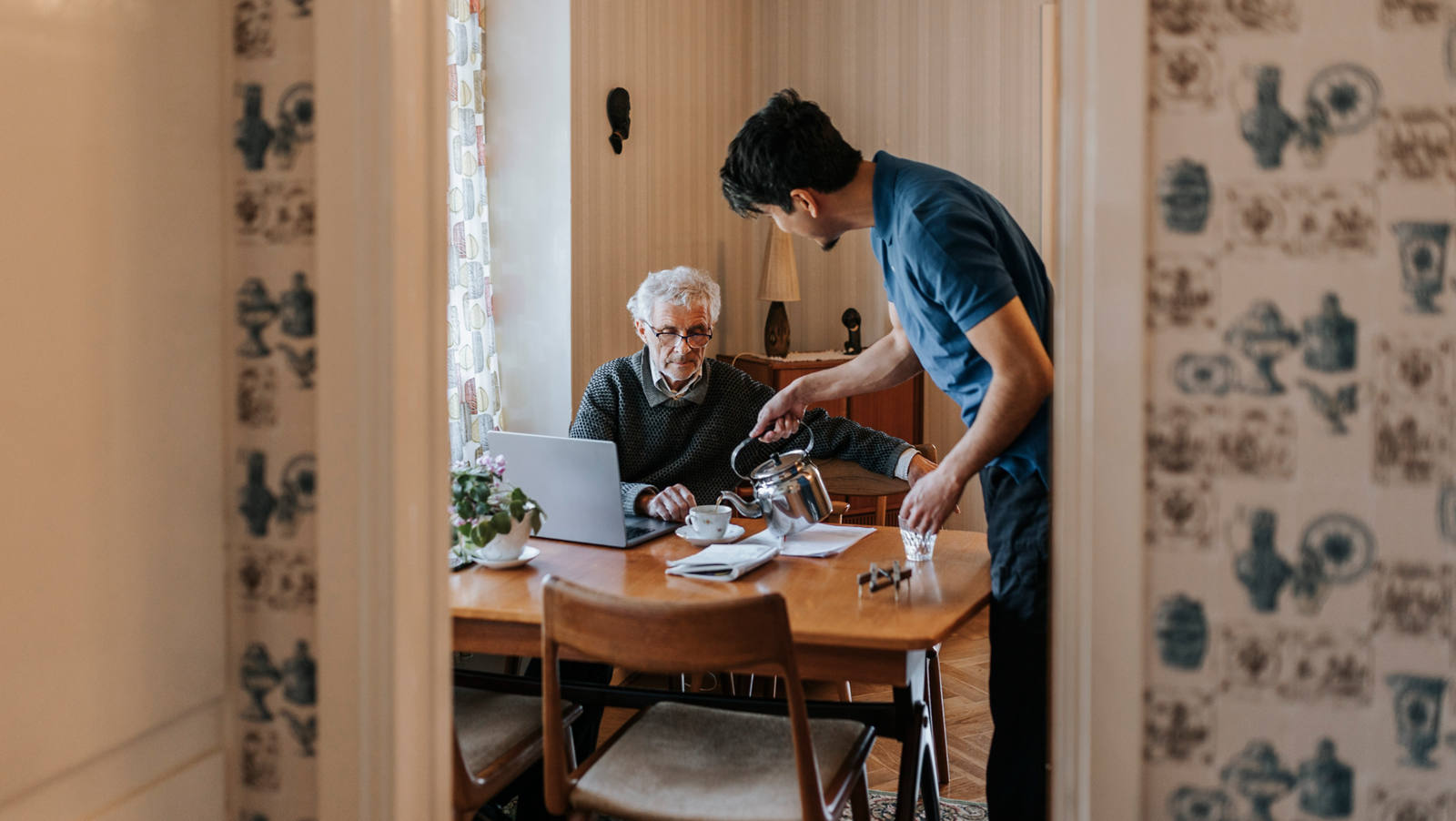 Ein junger Angehöriger hilft einem älteren Herrn zuhause und giesst ihm eine Tasse Tee ein.