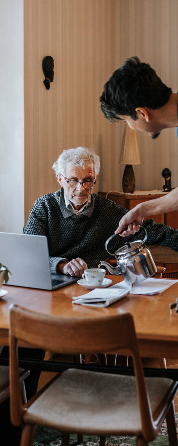 Ein junger Angehöriger hilft einem älteren Herrn zuhause und giesst ihm eine Tasse Tee ein.