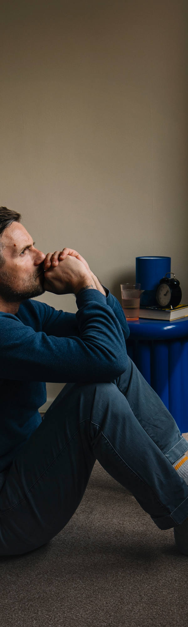 A man in pajamas sits on the floor in front of the bed and looks very thoughtfully and exhausted out of the window.
