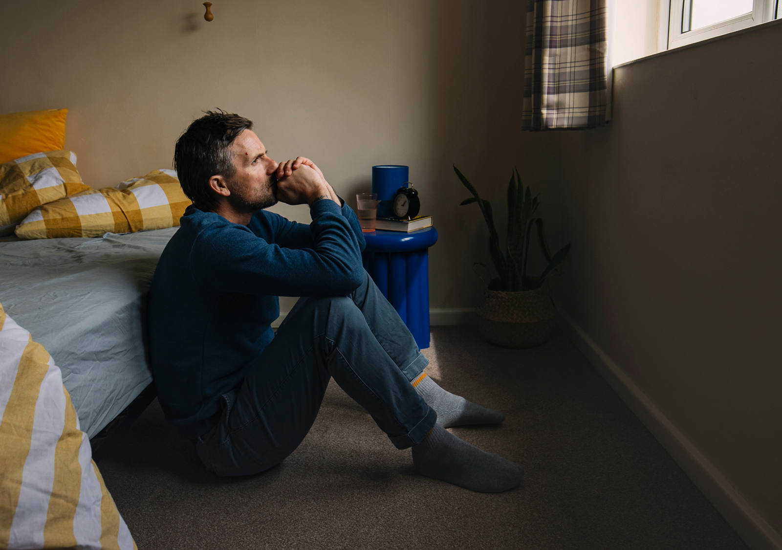 Ein Mann im Pyjama sitzt vor dem Bett am Boden und schaut sehr nachdenklich und erschöpft aus dem Fenster.