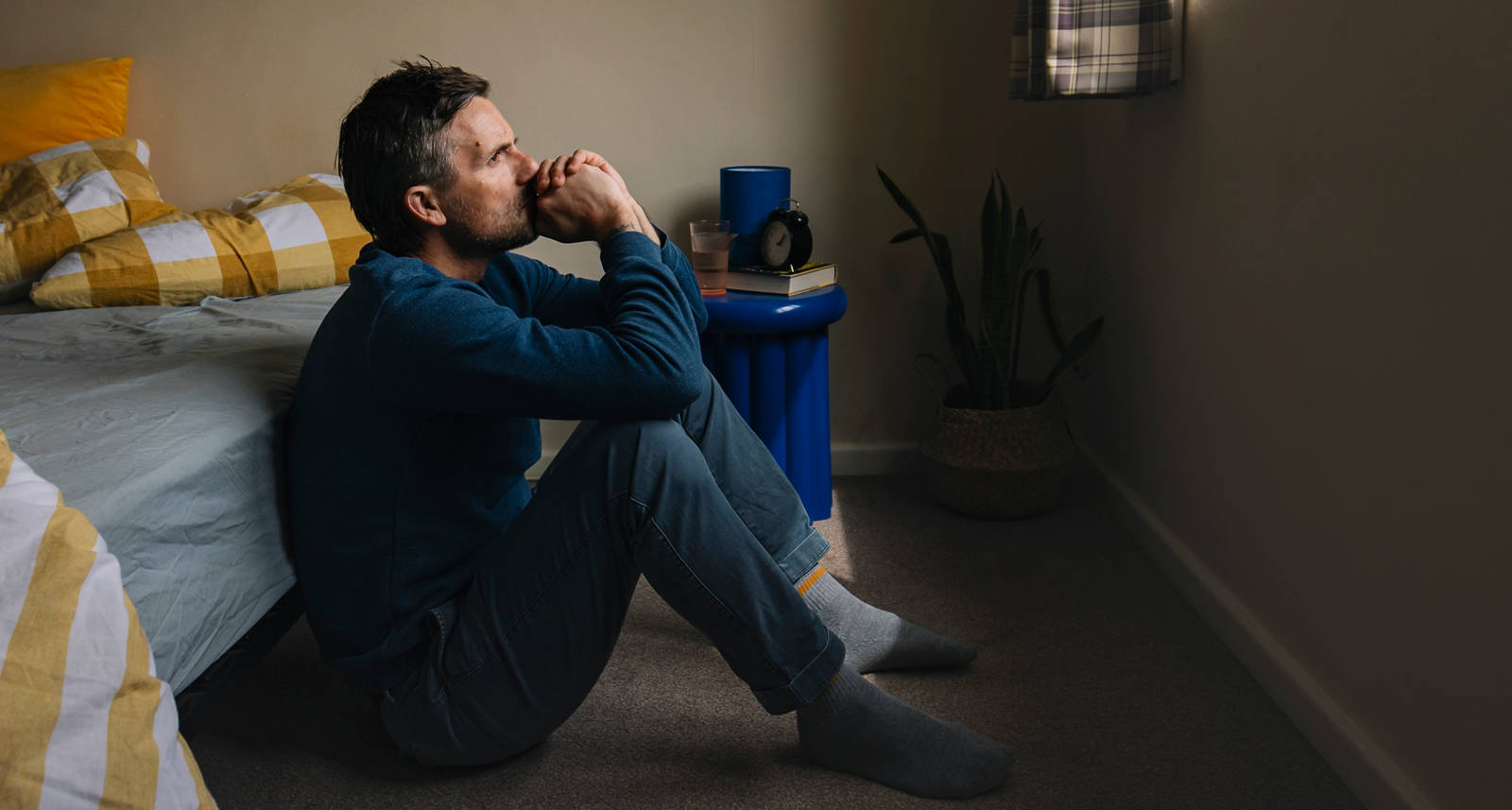 A man in pajamas sits on the floor in front of the bed and looks very thoughtfully and exhausted out of the window.