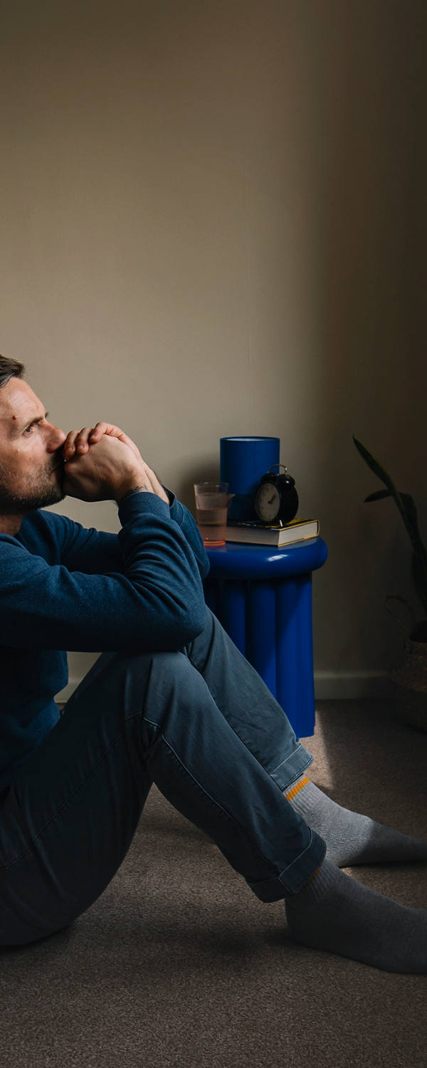 Un homme en pyjama est assis par terre devant le lit et regarde par la fenêtre très réfléchie et épuisé.