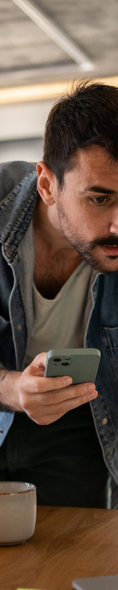 L’uomo con i baffi è appoggiato sopra il tavolo e confronta qualcosa sul laptop, tenendo in mano anche uno smartphone.