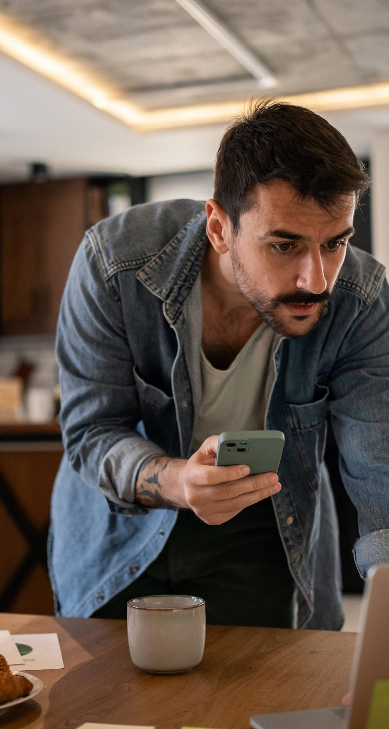 Man with a snout is leaning over the table and comparing something on the laptop while holding a smartphone in his hand.