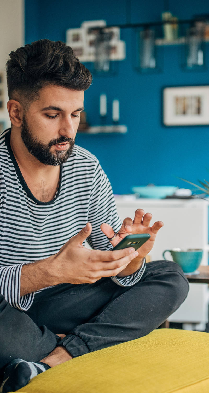 Young man sits comfortably on the armchair in the living room typing on the smartphone.