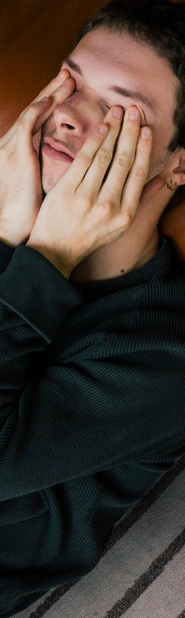 A man lies exhausted on the floor leaning against the sofa while rubbing his eyes.