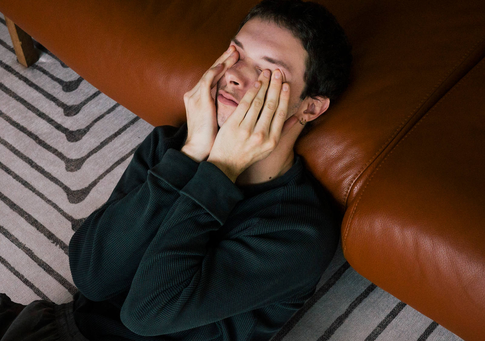Un homme est allongé par terre, épuisé, appuyé contre le canapé en se frottant les yeux.