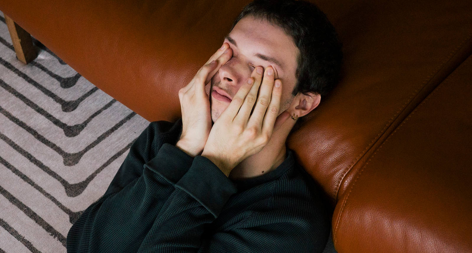 Ein Mann liegt erschöpft am Boden und lehnt gegen das Sofa, während er sich die Augen reibt.