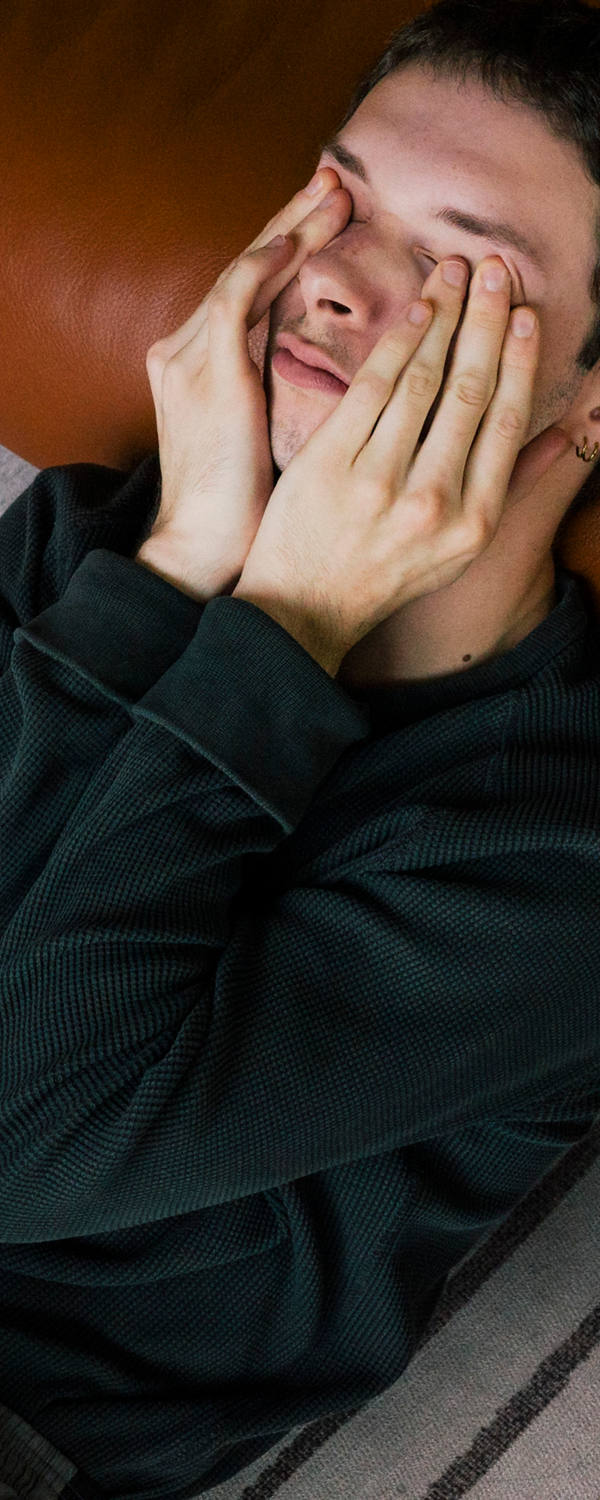A man lies exhausted on the floor leaning against the sofa while rubbing his eyes.