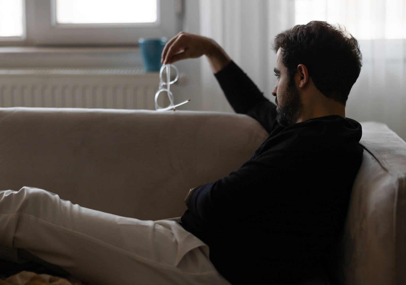 Un homme est assis sur le canapé, les jambes tendues, regardant d’un air pensif les lunettes qu’il tient dans sa main.