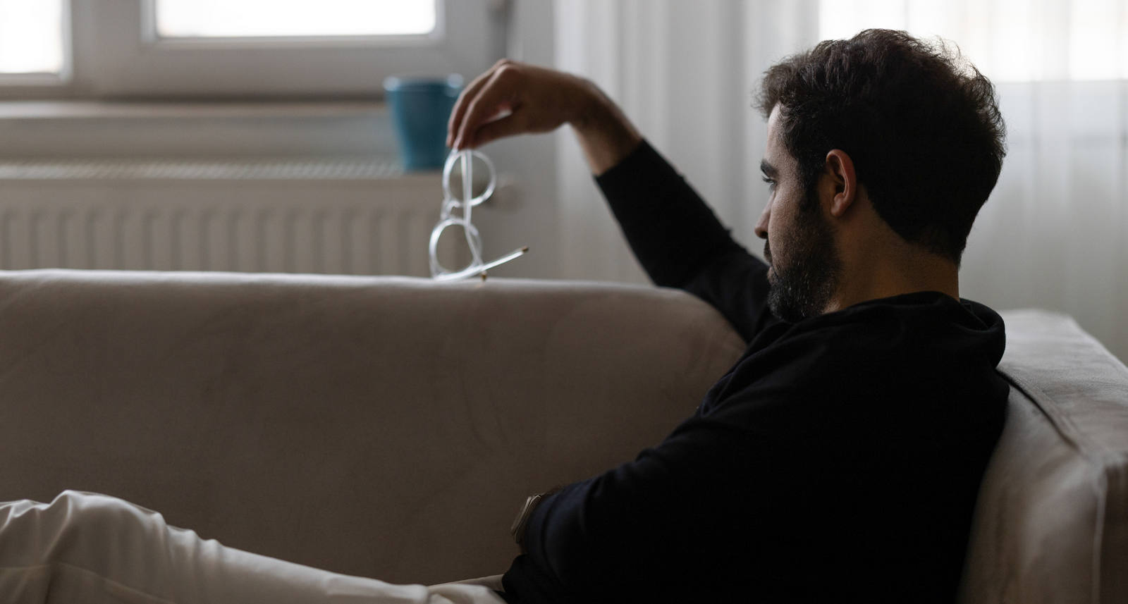 Ein Mann sitzt mit ausgestreckten Beinen auf dem Sofa und schaut nachdenklich auf die Brille, die er in seiner Hand hält.